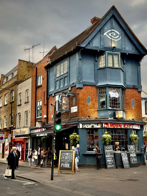 A lively street scene shows a historic corner building with a distinctive blue wooden facade and brickwork, housing a pub on the ground floor. The pub's exterior is decorated with hanging flower baske