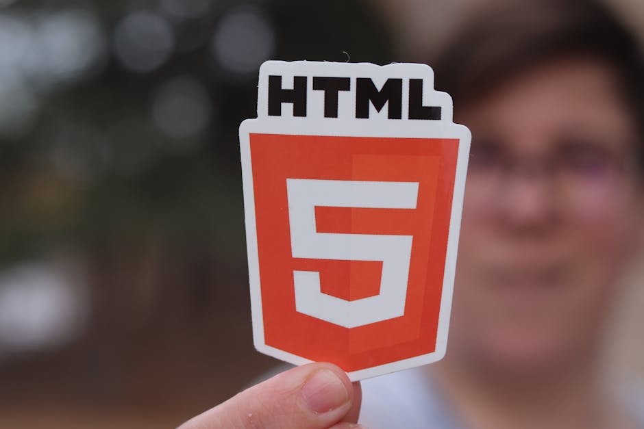 A person holding a small sign featuring the HTML5 logo with a bright red-orange shield and a white number 5 in the center; the background is blurred with hints of greenery, suggesting an outdoor setti